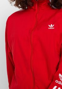 Röd Adidasjacka med hög krage, hel dragkedja och vitt logotyp på vänster bröst och ärm. Slät textur och lätt design.