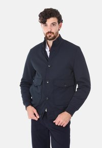 Giacca della marina con colletto alto, quattro tasche frontali e chiusura con bottoni. Tessuto liscio con design strutturato e polsini a coste.