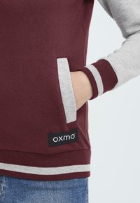 Mörkröd sweatshirt med grå ärmar, försedd med en sidficka, ribbade manschetter och en randig nederkant. Svart etikett med "oxmo"-logotypen nederst.