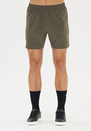 Homme portant un short de sport vert olive, des chaussettes noires mi-mollet et des baskets basses noires sur fond blanc.