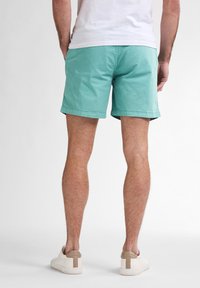 Shorts teal clair en coton, présentant un devant plat, des poches latérales et un ourlet soigné. Portés avec des baskets blanches et peu de détails.