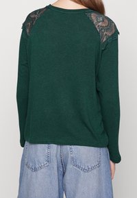 Dark green long-sleeve top with lace accents on shoulders, featuring a relaxed fit and a rounded hem. Paired with light blue denim shorts.