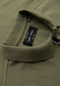 Olivgrön polo t-shirt med mjuk textur, med krage, sidoseam och en svart tvättlapp med "OMBRE EXTREMO" och storleksinformation.