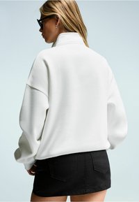 Felpa bianca oversize con colletto alto, presenta spalle scese e polsini a costine, abbinata a una mini gonna in denim nera.