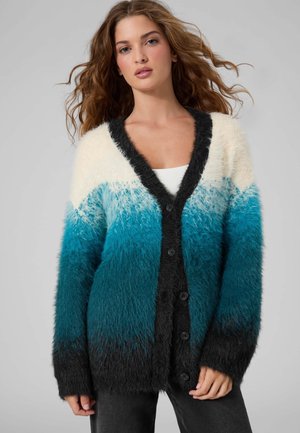 Femme aux cheveux bouclés porte un cardigan duveteux blanc, bleu et noir boutonné, un pantalon noir à jambes larges et des chaussures noires à talons, debout devant un fond gris.