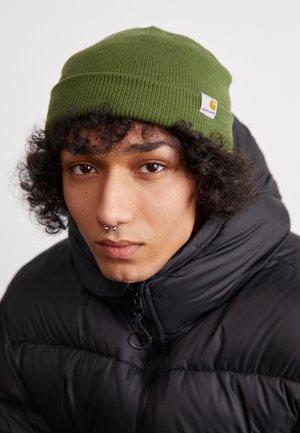 Gorro de punto verde con tela texturizada y una pequeña etiqueta de logo blanca y naranja, llevado con una chaqueta negra acolchada.