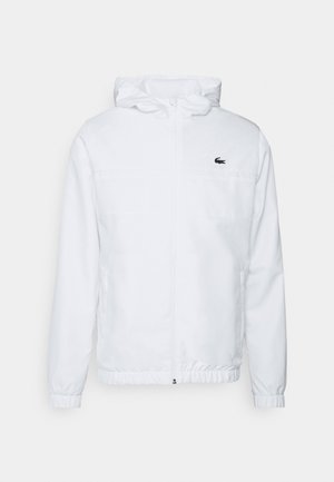 Valge kapuutsiga jakk, millel on elastsed mansetid ja stringiga hem. Rinnal on must logo ning külgedel kaks taskut, millel on sile tekstuur.