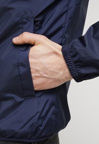 Marinblå vindjacka med elastiska ärmslut och en sidoficka. Slät, lätt material med en åtsittande form och rynkad nederkant.