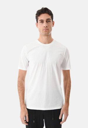 Uomo in piedi di fronte, che indossa una maglietta bianca a maniche corte semplice e pantaloni neri su uno sfondo grigio chiaro.