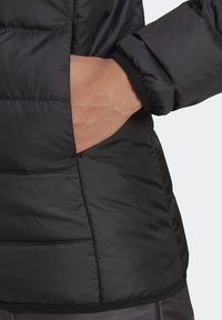 Černá péřová bunda má prošívanou texturu, elastické manžety a boční kapsu, přičemž je částečně vidět ruka. Materiál vypadá leskle a lehce.