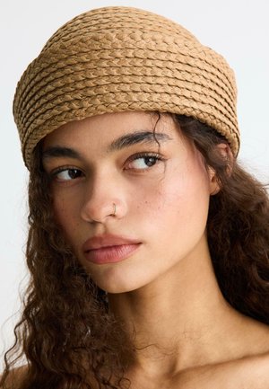 Mladá žena s kučeravými tmavými vlasmi, ktorá má na sebe tkaný béžový klobúk, pozerá sa do boku s malým nosným krúžkom na svetlom pozadí.