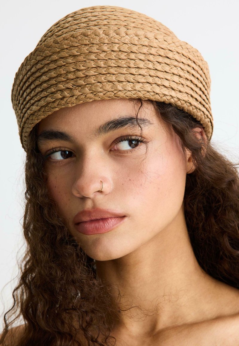 Jeune femme aux cheveux bouclés et foncés, portant un chapeau tressé couleur fauve, regardant sur le côté avec un petit anneau au nez sur un fond clair.