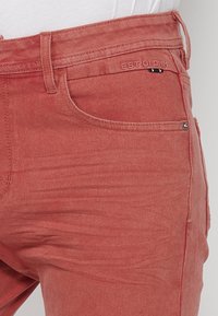 Jeans de mezclilla roja con corte ajustado, que cuentan con un bolsillo frontal, pasadores para cinturón y un logotipo en relieve cerca de la cintura. Textura suave.