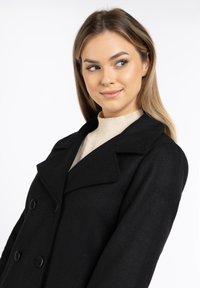 Black wool peacoat with double-breasted buttons, wide lapels, and a textured finish. Features embroidery on the sleeve and a cream turtleneck underneath.