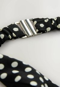 Bandeau en tissu noir à pois blancs avec une fermeture à clip en métal argenté sur fond clair.