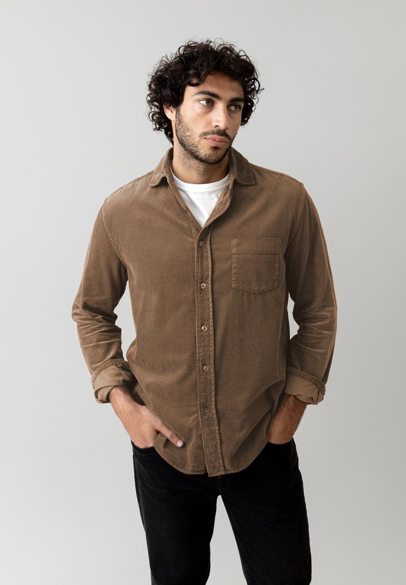 Camicia di cotone a coste marrone con bottoni frontali, taschino sul petto e maniche arrotolate. Indossata con una t-shirt bianca a collo rotondo sotto.