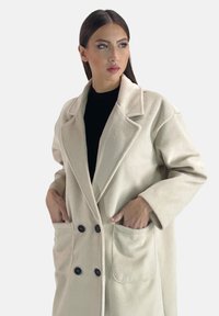 Dubbelknäppt, gräddfärgad kappa i mjukt material med stora fickor och svarta knappar. Lös passform med slagkrage.