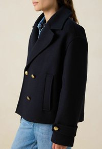 Manteau en laine navy à double boutonnage, avec des boutons dorés, un large col et une poche latérale, associé à un jean bleu clair.