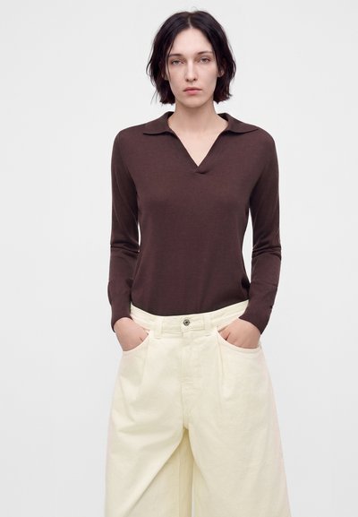 Jeune femme aux cheveux courts et foncés, portant un haut marron à manches longues et un pantalon blanc ample, les mains dans les poches, debout devant un fond uni.