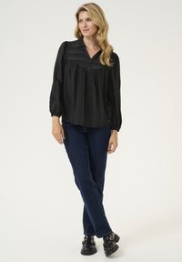 Melnas, teksturētas blūzes ar apkakli, garām piedurknēm un plisētu dizainu. Saskaņota ar tumši zilu džinsu biksēm un melnām kurpēm.