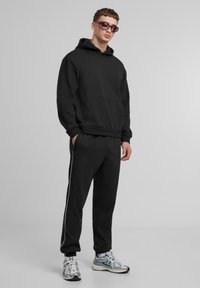 Schwarzer Kapuzenpullover mit Fronttasche und Kordelzug, kombiniert mit schwarzen Jogginghosen mit weißen Seitenstreifen. Das Modell trägt graue Sneaker und Sonnenbrille.