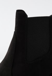 Botín de ante negro con panel lateral elástico, puntera redondeada, detalles de costura fina y textura suave.