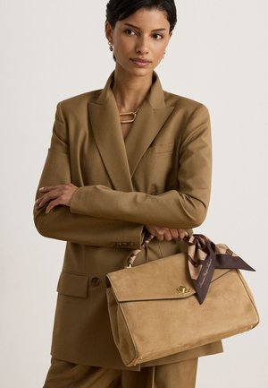 Vrouw in een bruin pak houdt een beige suède handtas vast met een patroon sjaal die aan het handvat vastzit tegen een effen achtergrond.