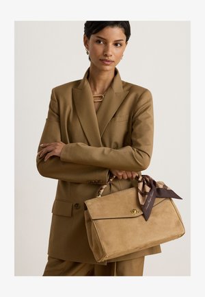 Femme portant un costume marron tient un sac à main en daim beige avec un foulard à motifs attaché à la poignée, sur un fond uni.