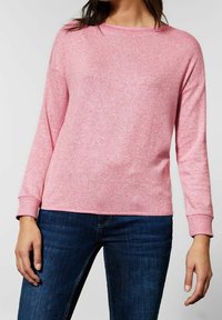 Jasnoróżowy sweter z długim rękawem wykonany z miękkiej, teksturowanej tkaniny, z okrągłym dekoltem i ściągaczami na mankietach, w połączeniu z ciemnoniebieskimi dżinsami.
