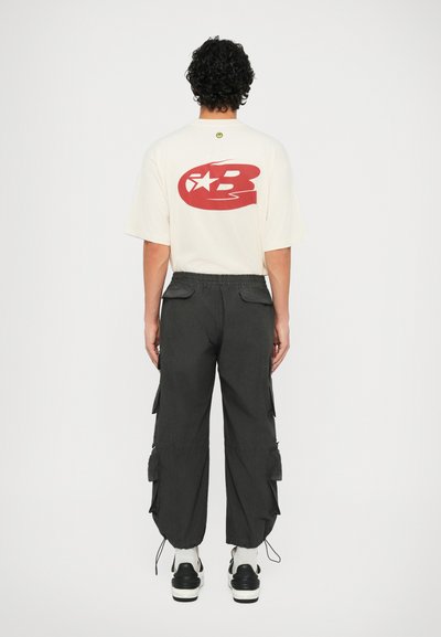 Κρεμ oversize μπλούζα με κόκκινη εκτύπωση στην πλάτη, συνδυασμένη με μαύρο cargo παντελόνι με πλαινές τσέπες και ελαστικές μανσέτες; μαύρα αθλητικά παπούτσια.