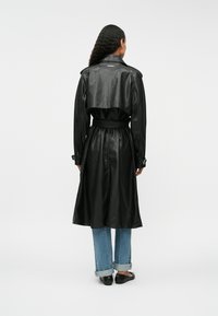 Czarny skórzany płaszcz trench z paskiem w talii, długimi rękawami i perforowanym wzorem. Modelka ma na sobie dżinsy z podwiniętymi nogawkami i baleriny.