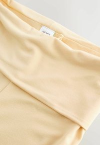 Veniv helekollane soonikkoes vöökoht, millel on pehme tekstuur ja nähtav valge silt, millel on kiri „NEXT“. Kangas tundub sile ja õmblustevaba.