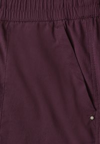 Gros plan sur un pantalon violet foncé avec une taille élastique, détail de poche latérale et petit bouton métallique près du bord de la poche.