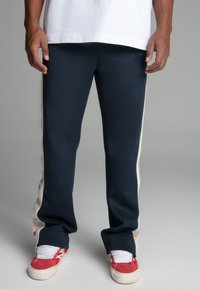 Navy tracksuit pants with cream side panels. Made of smooth fabric, featuring an embroidered logo on the left thigh and tapered fit.
