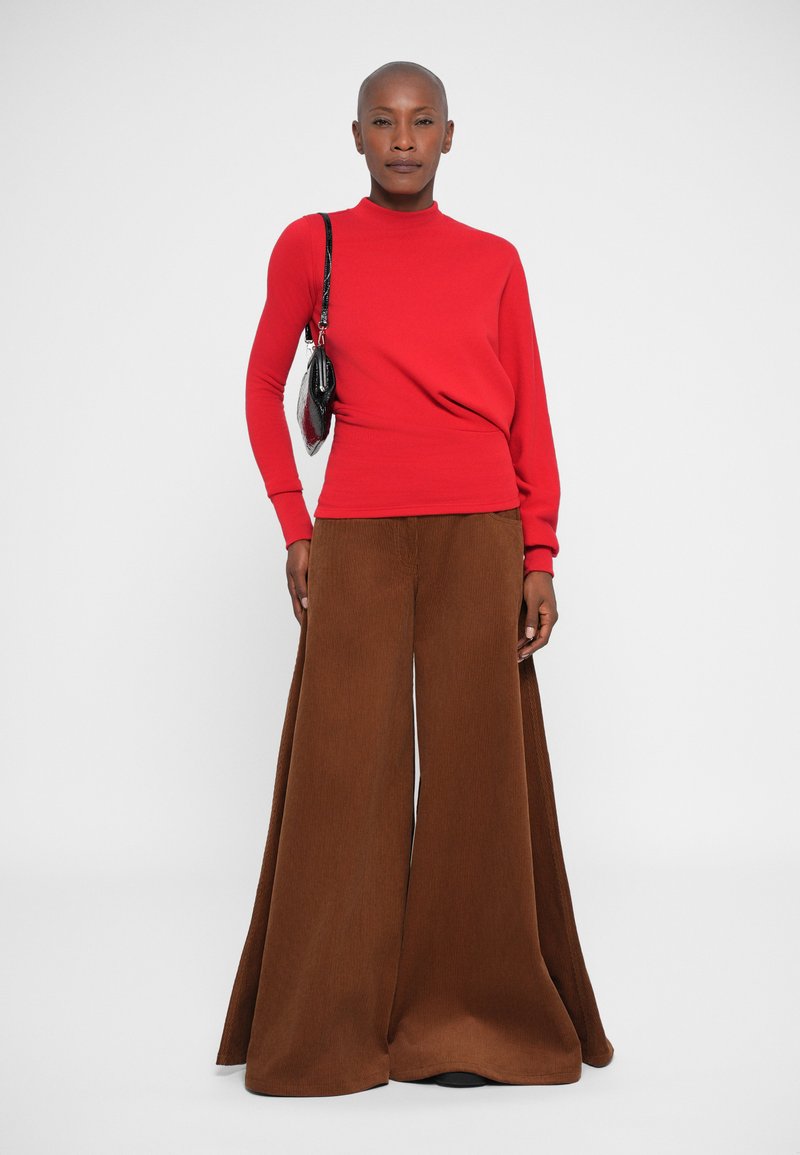 Haut rouge à manches longues avec un col haut, associé à un pantalon en velours côtelé marron à jambes larges. Le modèle tient un petit sac à main noir.