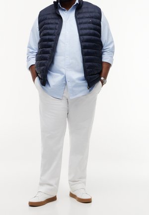 Mann trägt weiße Hose, hellblaues Hemd mit Knöpfen, marineblaue gesteppte Weste, weiße Sneakers und eine Armbanduhr, steht mit Händen in den Taschen.