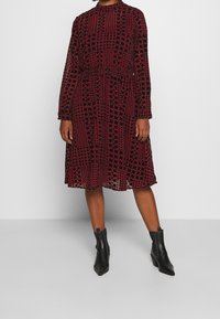 Röd och svart mönstrad klänning med långa ärmar, åtsittande midja och flödande kjol. Bärs med svarta anklar stövlar. Strukturerad, halvgennomsiktig kvalitet.