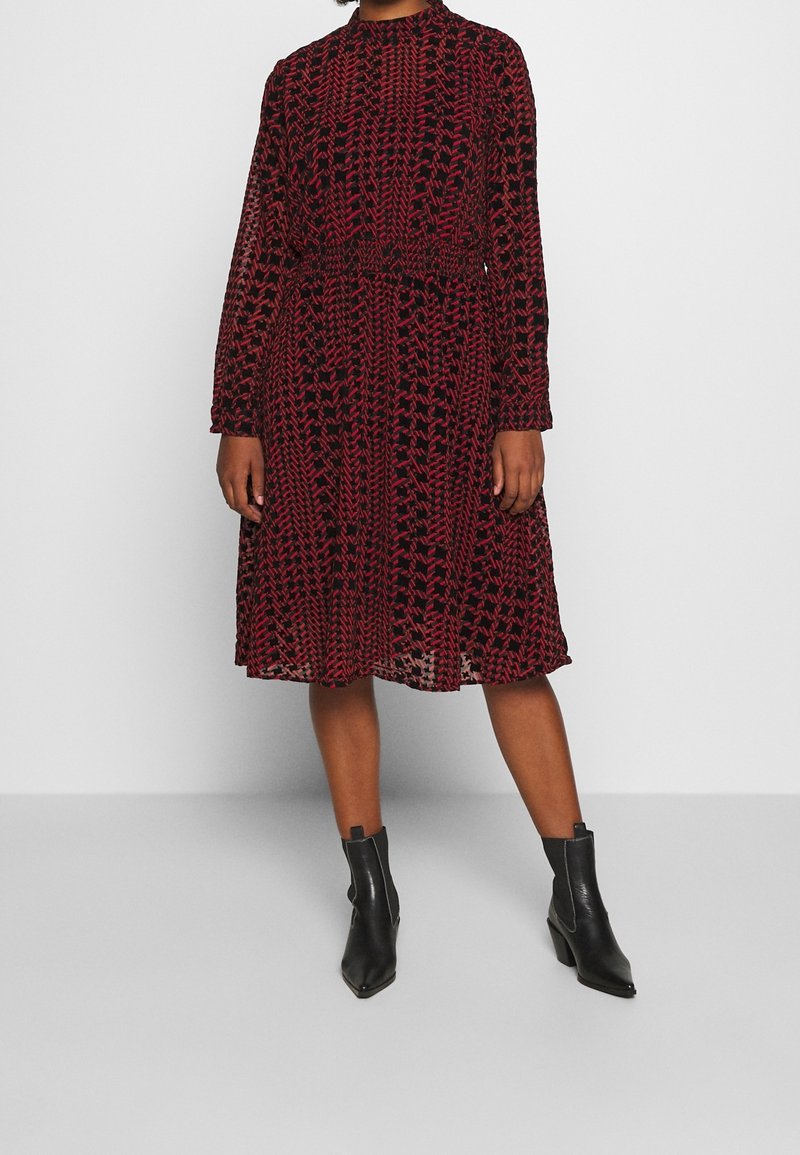 Röd och svart mönstrad klänning med långa ärmar, åtsittande midja och flödande kjol. Bärs med svarta anklar stövlar. Strukturerad, halvgennomsiktig kvalitet.