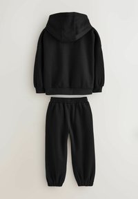 Černá bavlněná mikina s kapucí a žebrovanými manžetami, v kombinaci s volnými kalhotami stejné barvy, které mají elastický pas a stažené kotníky.