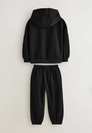 Melna kokvilnas kapuce ar kapuci un ribotām piedurknēm, apvienota ar atbilstošām brīvām biksēm ar elastīgu jostu un savilktiem potīšu galiem.