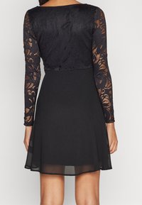 Kvinna i en svart klänning med genomskinliga spetslånga ärmar och en enfärgad kjol, sedd bakifrån mot en ljus neutral bakgrund.