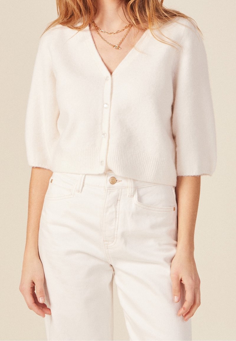 Cardigan bianco, corto, con scollo a V, maniche corte a sbuffo e bottoni perla. Abbinato a jeans chiari a vita alta.