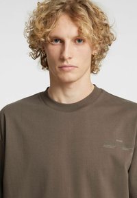 Rjava bombažna majica z okroglim ovratnikom, na sprednji strani z nevsiljivimi grafičnimi napisi in gladko, enobarvno teksturo.