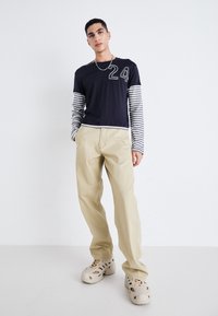 Chemise noire à manches courtes avec des manches longues rayées blanches, arborant un grand design '24' ; pantalons beige à jambes larges ; sabots beige.