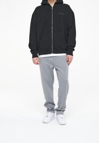 Schwarze Zip-Up-Hoodie mit einer Fronttasche, kombiniert mit hellgrauen Jogginghosen mit Kordelzug, getragen mit weißen Sportschuhen.