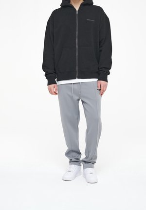Schwarze Zip-Up-Hoodie mit einer Fronttasche, kombiniert mit hellgrauen Jogginghosen mit Kordelzug, getragen mit weißen Sportschuhen.