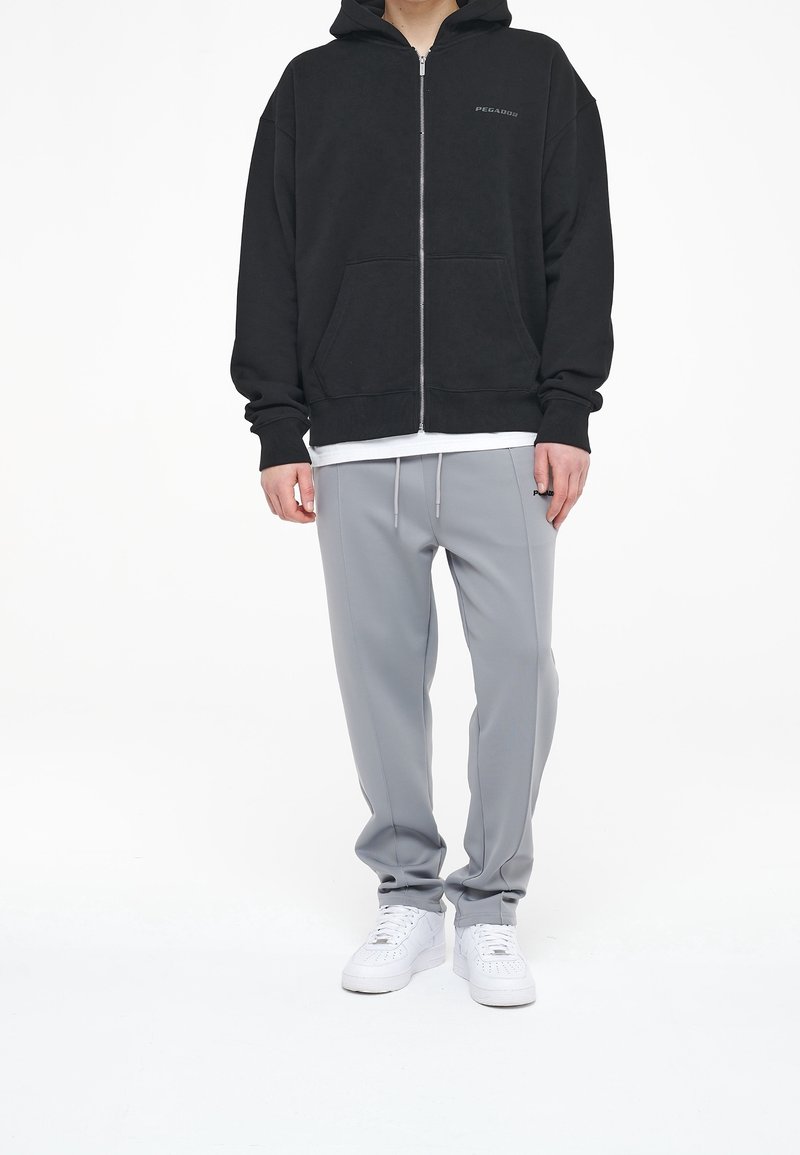 Schwarze Zip-Up-Hoodie mit einer Fronttasche, kombiniert mit hellgrauen Jogginghosen mit Kordelzug, getragen mit weißen Sportschuhen.