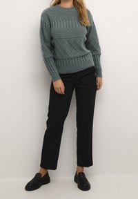 Maglione lavorato a maglia in teal opaco con motivi a diamante e verticali testurizzati. Abbinato a pantaloni neri su misura e scarpe slip-on nere.