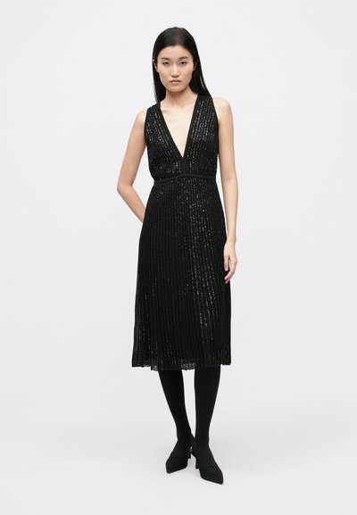 Fekete midi ruha mély V-nyakkivágással, függőleges flitterekkel díszítve és szabott derékkal, fekete harisnyával és magassarkúval párosítva.