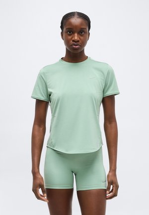 Vrouw met gevlochten haar, gekleed in een lichtgroen sportief T-shirt met korte mouwen en bijpassende shorts, staand tegen een effen achtergrond.